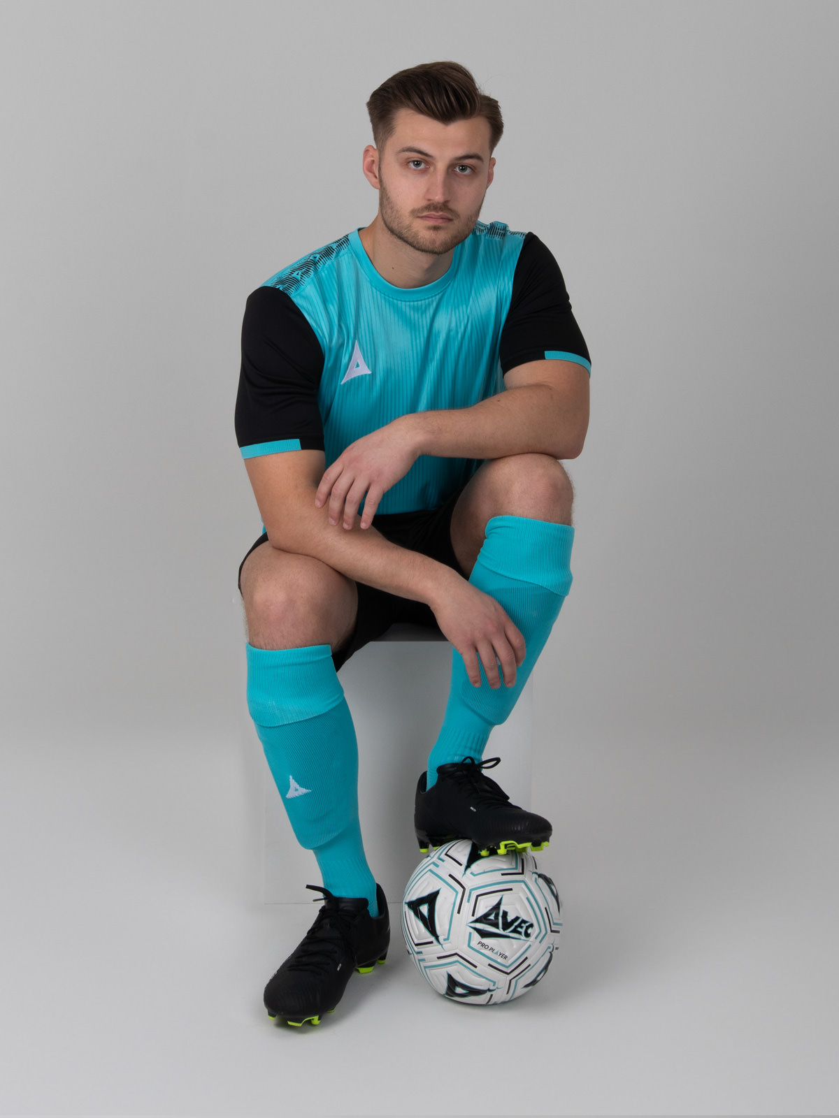 a model is sitting down posing in a light blue football shirt with black sleeves, black shorts and pale blue socks
