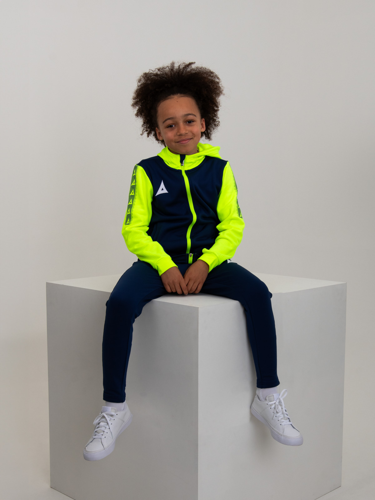 a young boy sitting on a box wearing a navy and yellow hoody and matching navy jogging bottoms