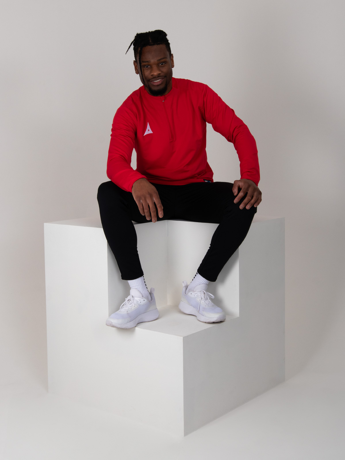 the model is sitting down on a box wearing a red jumper with a zip and black joggers.