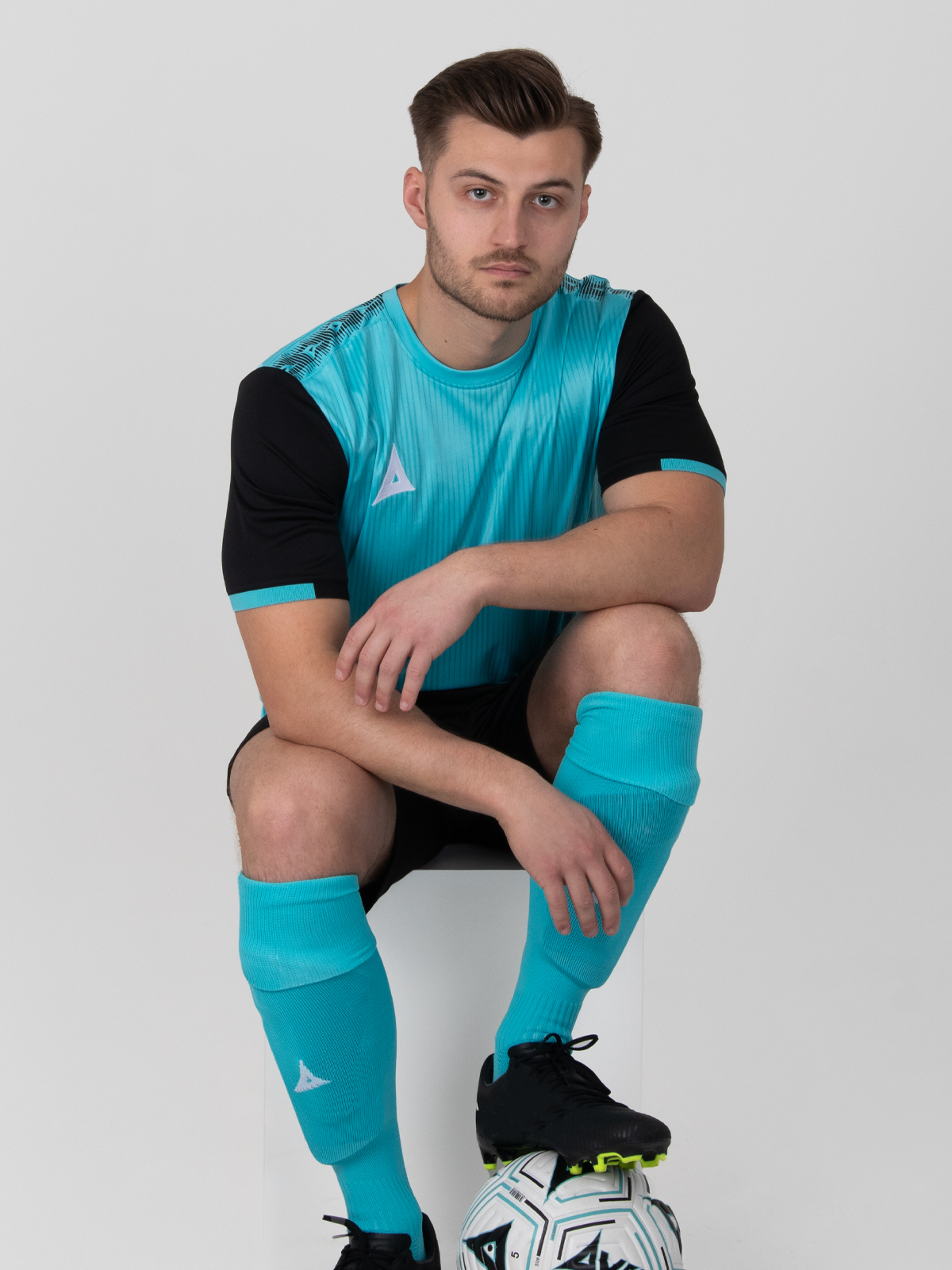 a model is sitting down posing in a light blue football shirt with black sleeves, black shorts and pale blue socks