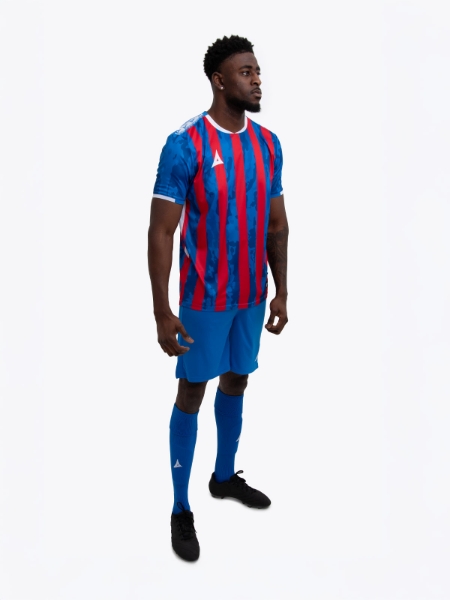 A model standing up wearing a red and royal blue football shirt, blue Shorts and blue Football Socks.