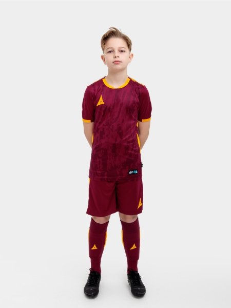 A kid standing up wearing a claret / maroon football shirt, matching claret Shorts and claret Football Socks.