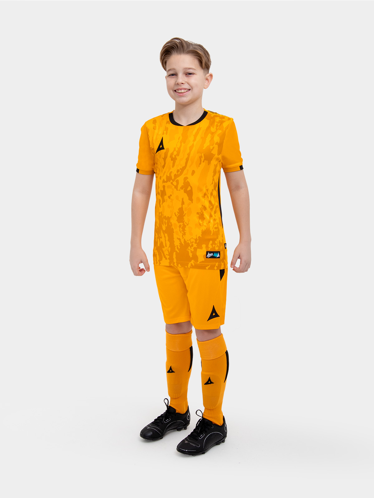 a kid standing up wearing an amber / black football shirt, matching amber shorts and amber football socks.