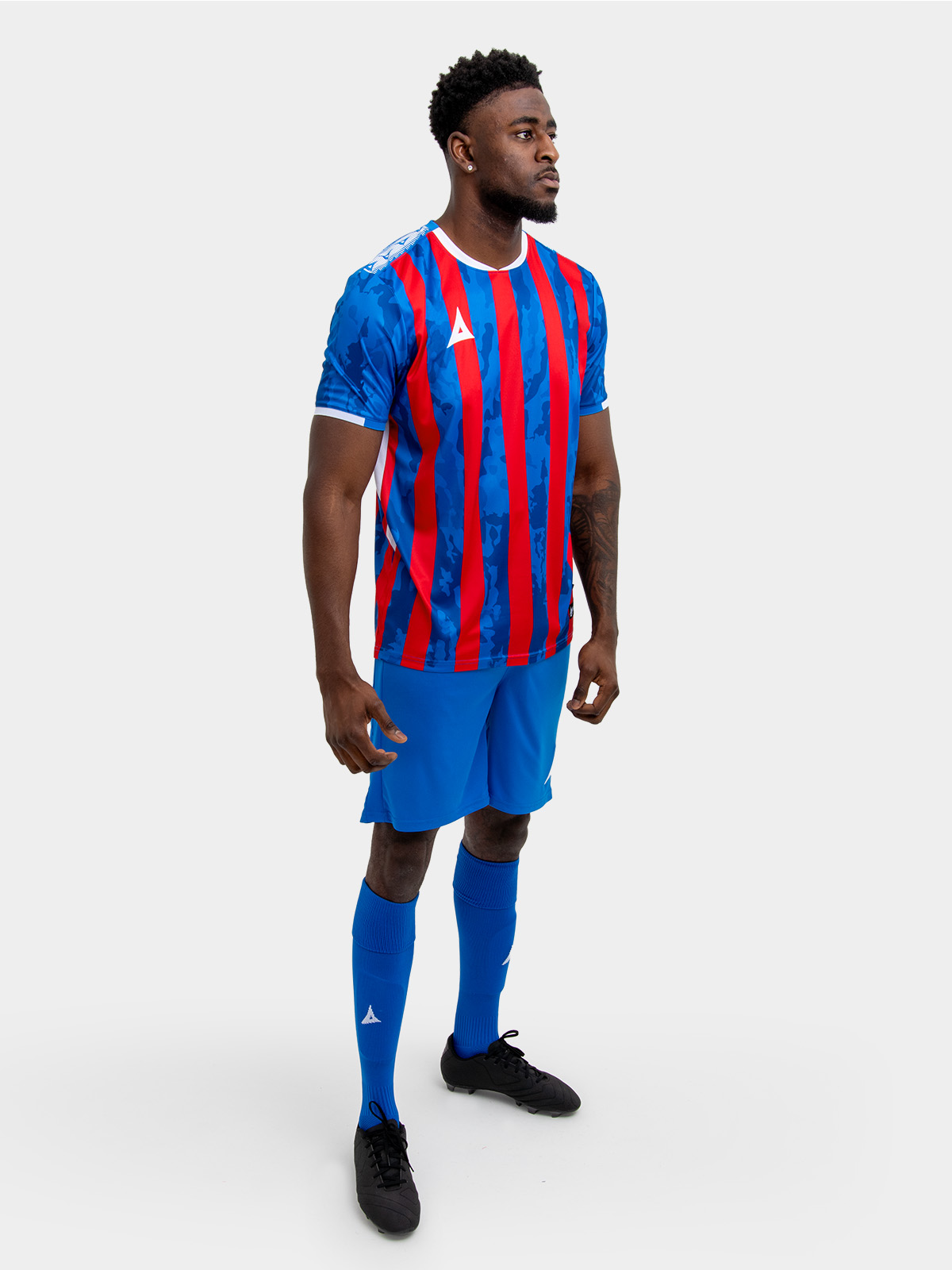 a model standing up wearing a red and royal blue football shirt, blue shorts and blue football socks.