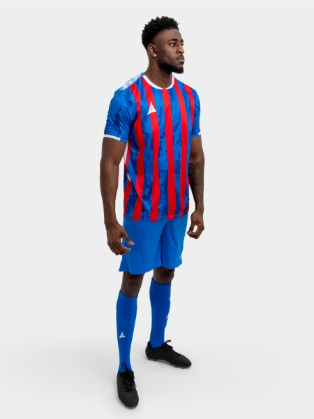 A model standing up wearing a red and royal blue football shirt, blue Shorts and blue Football Socks.