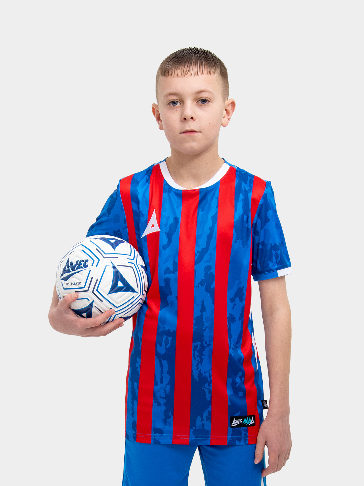 	a kid standing up wearing a red and royal blue football shirt and holding a royal blue and white football