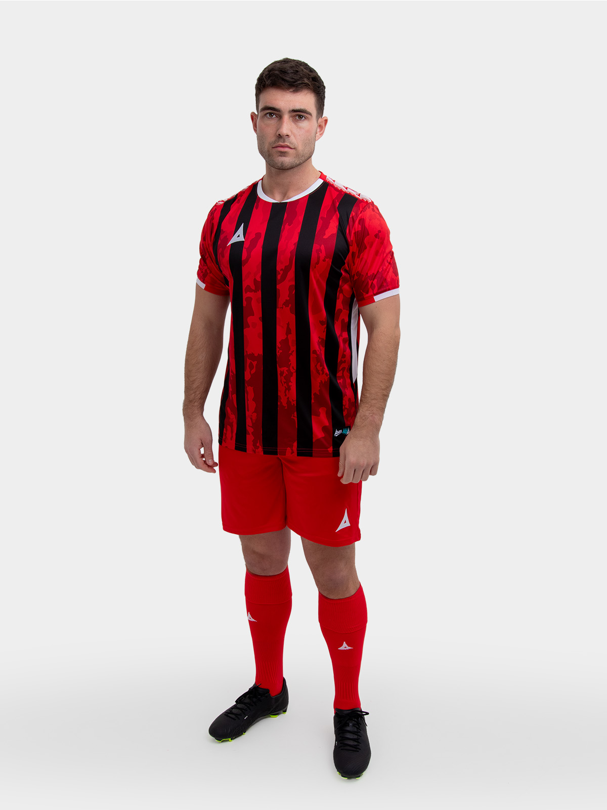 a model standing up wearing a red and black football shirt, red shorts and red football socks.
