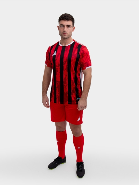 A model standing up wearing a red and black football shirt, red Shorts and red Football Socks.