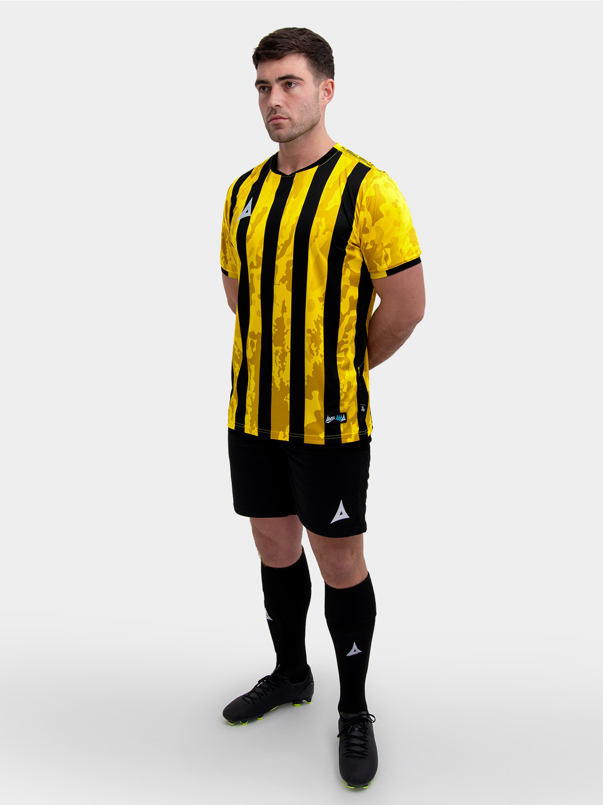 a model standing up wearing a yellow and black football shirt, yellow shorts and black football socks.