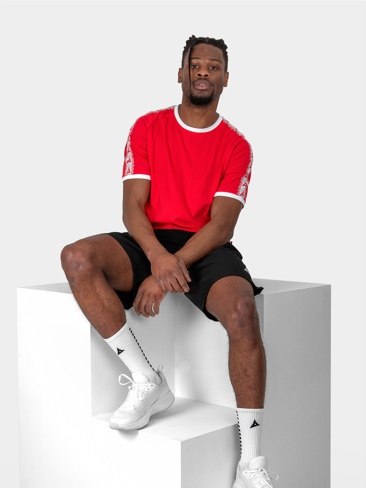 	a male model is sat wearing the avec evolve red t-shirt, and black avec focus 2 tech shorts and avec grip socks in white