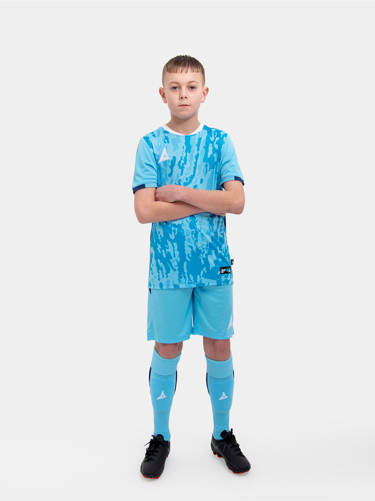 	a kid standing up wearing a sky / navy football shirt, matching shorts and football socks.
